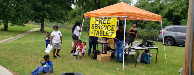 A group of individuals standing in a tent with a banner that reads Free Service plus Tablet
